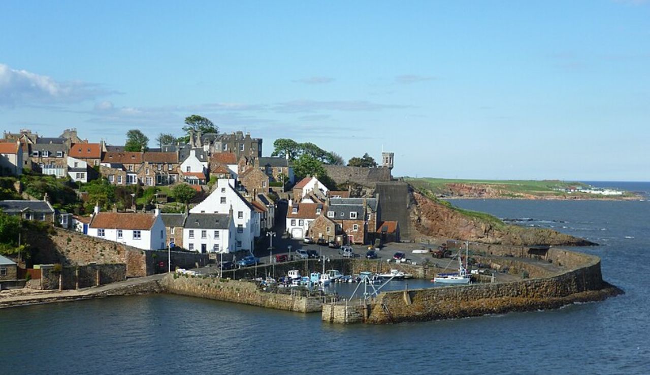 A charming coastal village with stone houses and a small harbor, surrounded by a curved seawall and set against the open sea and green cliffs.