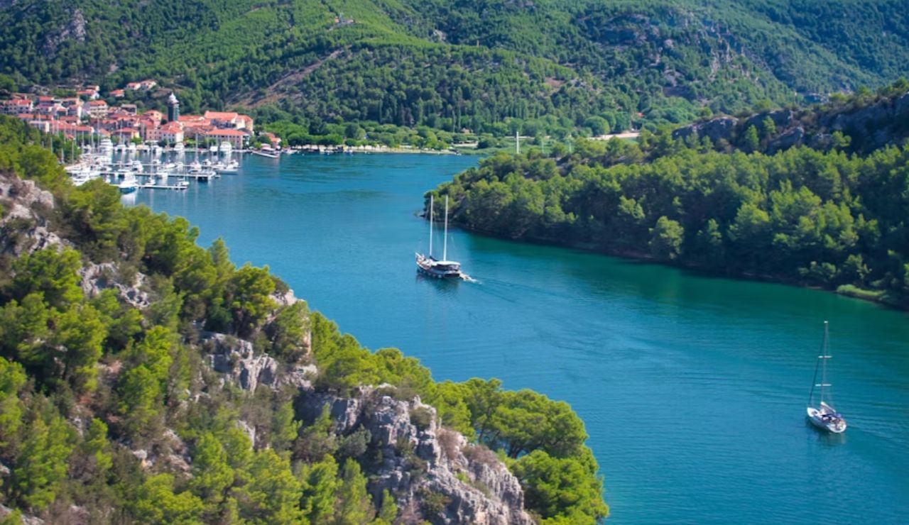 Sailboats cruising through a lush green river valley near a coastal village.