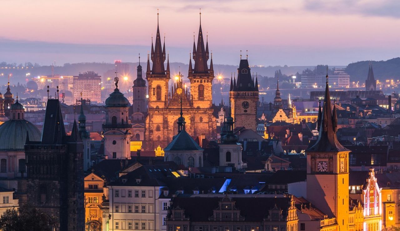 Evening view of Prague’s Old Town with the illuminated Church of Our Lady before Týn and historic towers against a soft purple sky.