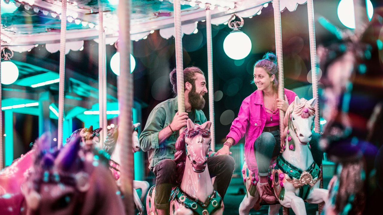 Couple riding colorful carousel horses at night, smiling and holding hands under bright lights.