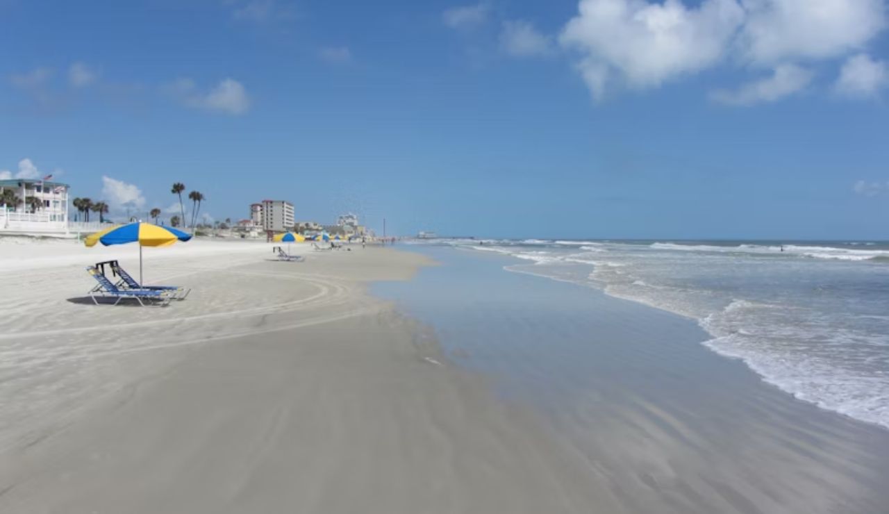 A quiet beach with yellow and blue umbrellas and chairs set up along the shoreline, with gentle waves and a few buildings in the distance.
