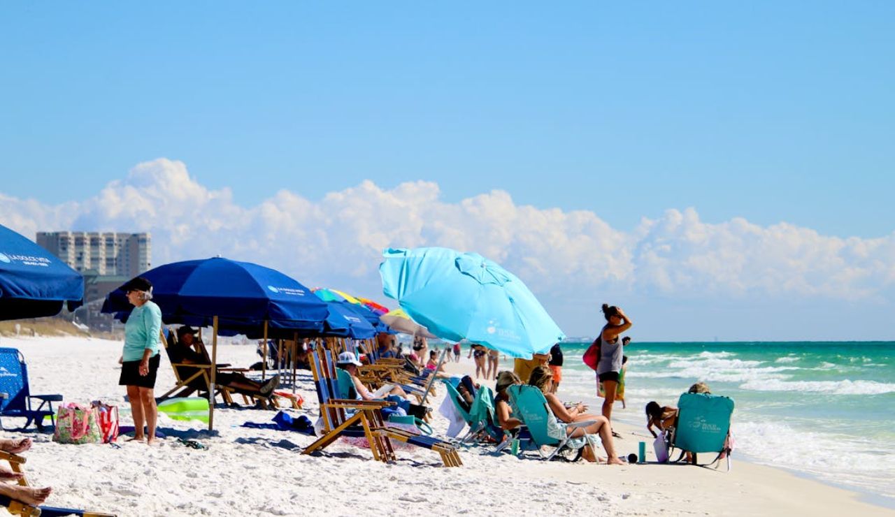 A lively beach scene with people relaxing under colorful umbrellas and lounging on beach chairs by the turquoise water.