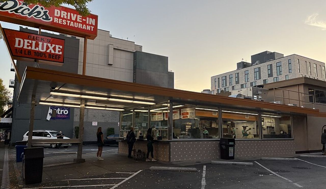 People wait in line at Dick's Drive-In Restaurant in Seattle at dusk, with the retro-style building and iconic sign lit under a twilight sky.