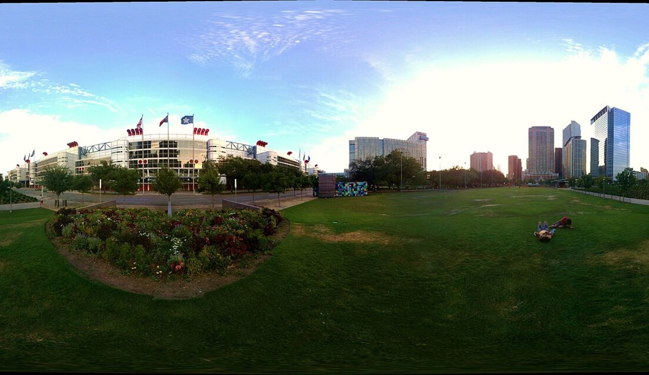 Wide-angle view of a city park with flowerbeds, open grassy space, and a modern building with flags, set against a backdrop of skyscrapers at sunset.