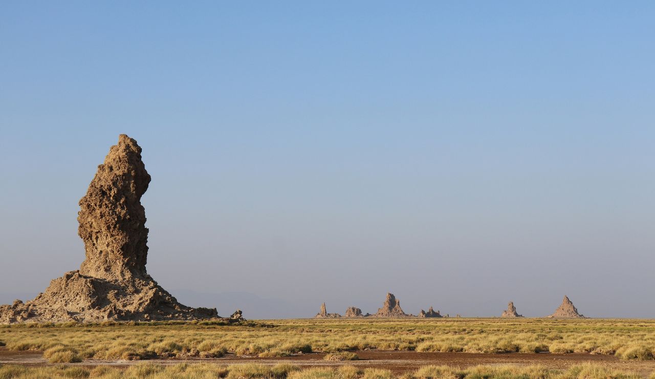 Tall, jagged rock formation standing alone in a vast, dry desert landscape under a clear blue sky.