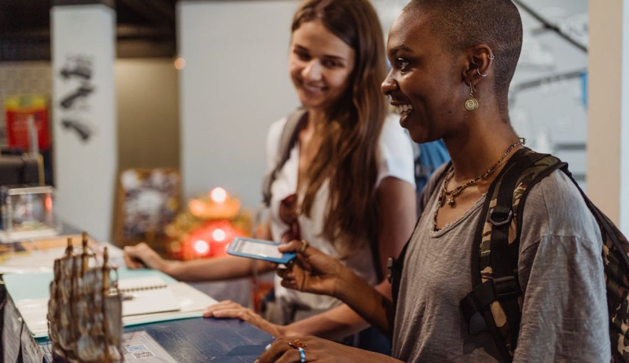 Two smiling women at a counter booking a cruise with phone and documents.