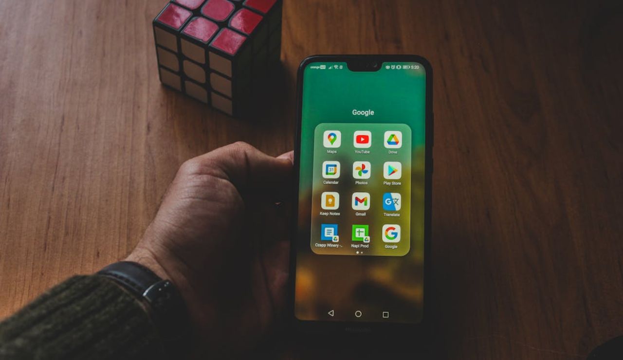 Hand holding a smartphone displaying Google apps on the screen, with a Rubik's cube on a wooden table in the background.