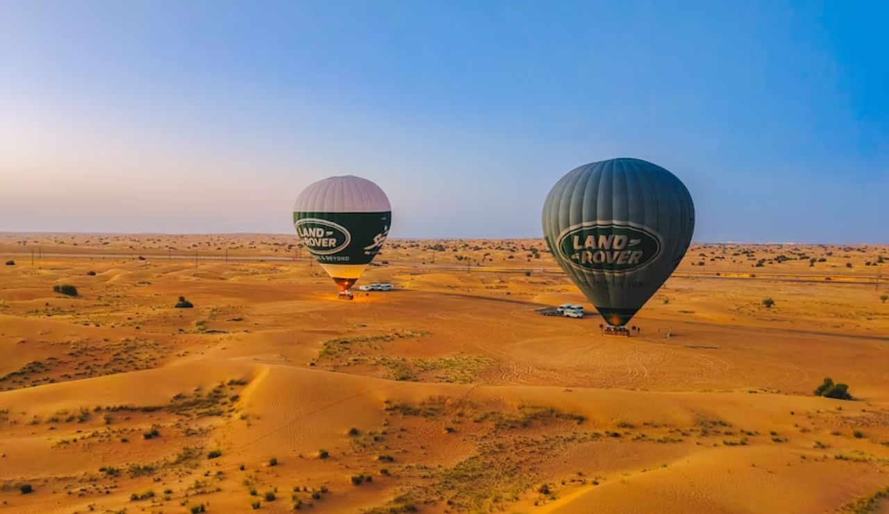 Two Land Rover-branded hot air balloons prepare for liftoff over the golden sand dunes of Dubai, UAE, under a clear blue sky.