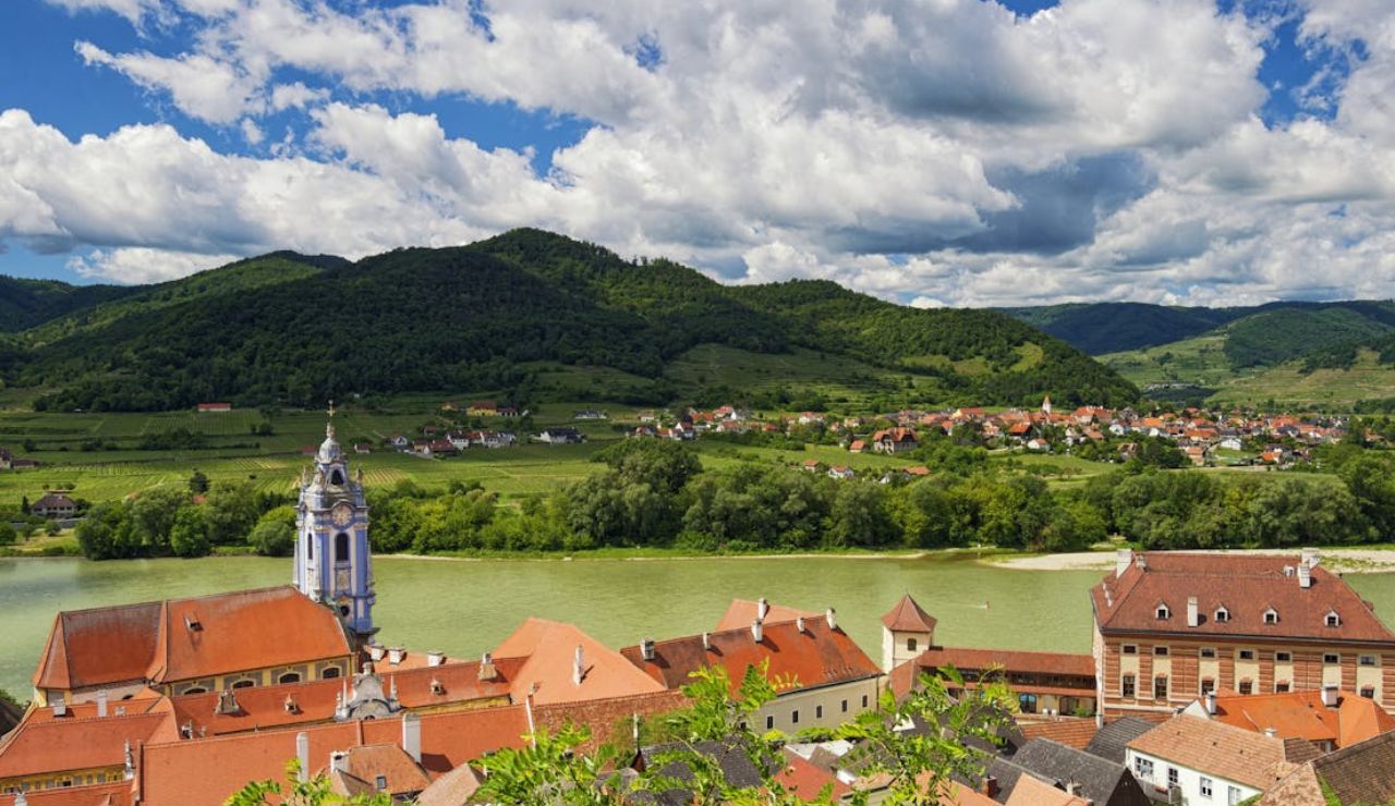 Scenic view of Dürnstein village in Austria with red-roofed buildings, a blue church tower, and green hills across the Danube River.