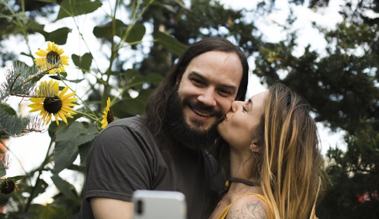 A smiling man takes a selfie with a woman who is kissing his cheek, surrounded by sunflowers and greenery.