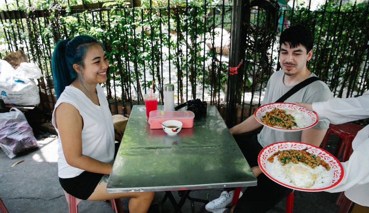Two people sitting at an outdoor metal table, smiling as a server brings plates of local rice and meat dishes to their table.