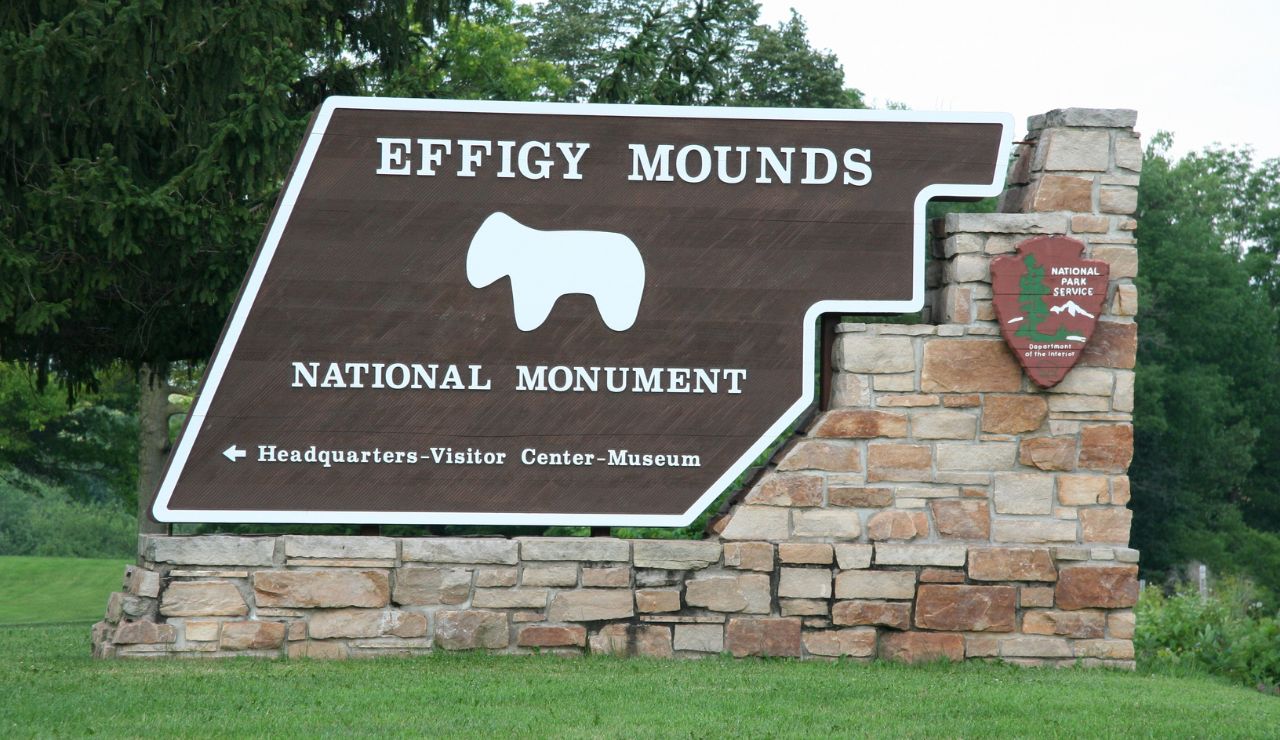 The Effigy Mounds National Monument entrance sign, featuring the National Park Service emblem, stands amid grass and trees.