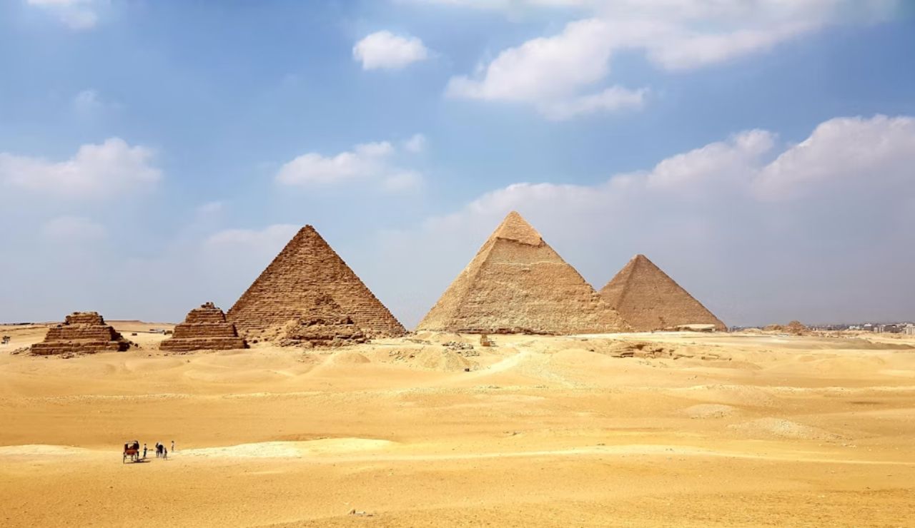 The Great Pyramids of Giza standing prominently on a sandy desert plain under a partly cloudy sky with tourists nearby.