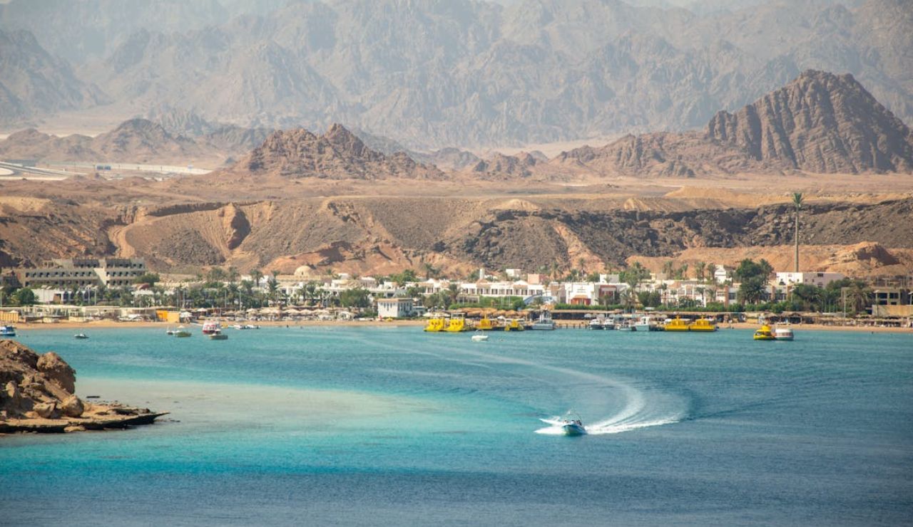 Speedboat cruising in turquoise waters near a desert coastal town with mountains.