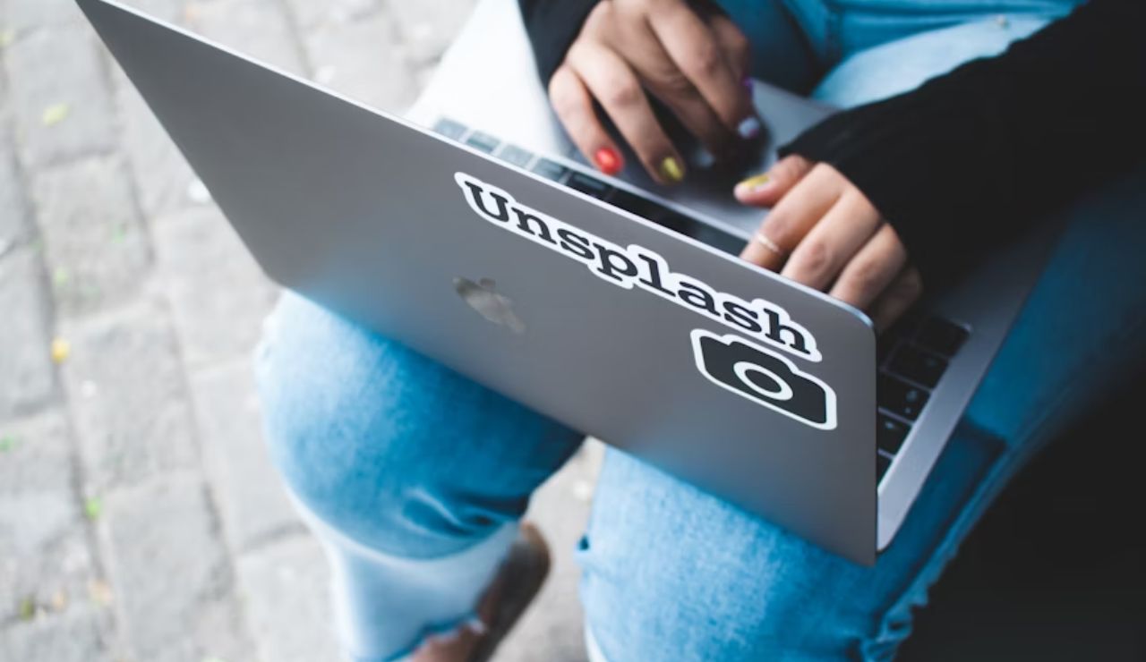 Close-up of a person using a laptop with an 'Unsplash' sticker, sitting outdoors in ripped jeans and colorful painted nails.