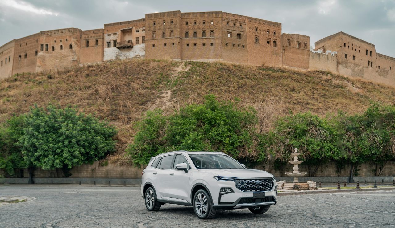 White SUV parked in front of the historic Erbil Citadel in Kurdistan, Iraq, with stone walls and greenery in the background.