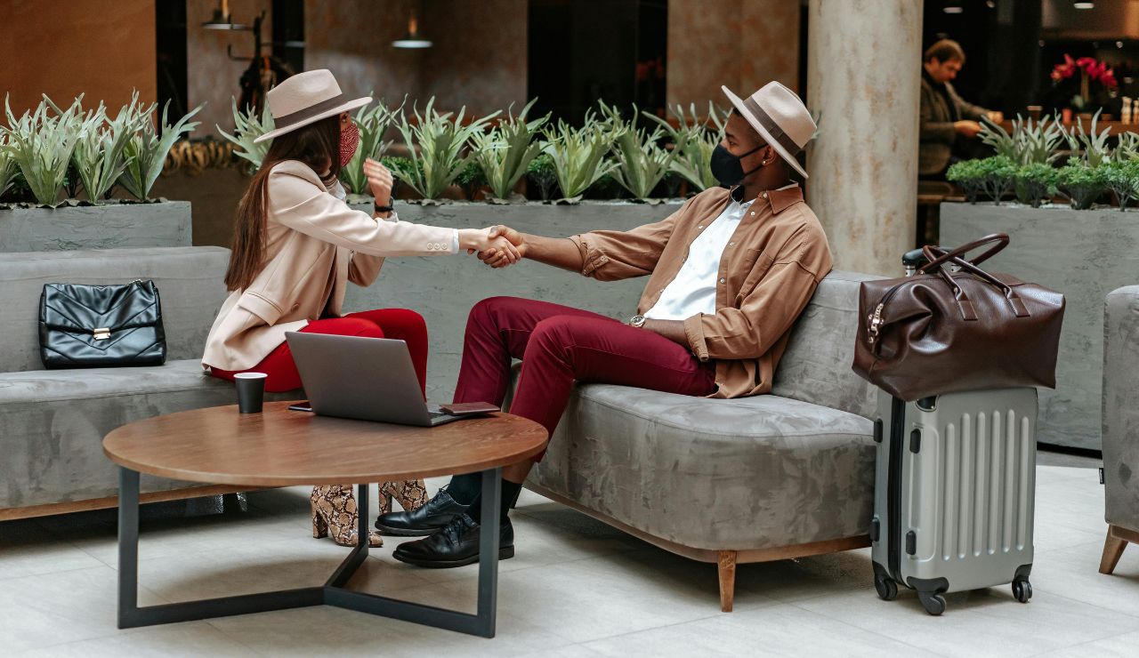 Two stylish people wearing hats and face masks shaking hands while sitting on lounge chairs in a modern indoor space, with luggage, a laptop, and coffee nearby.