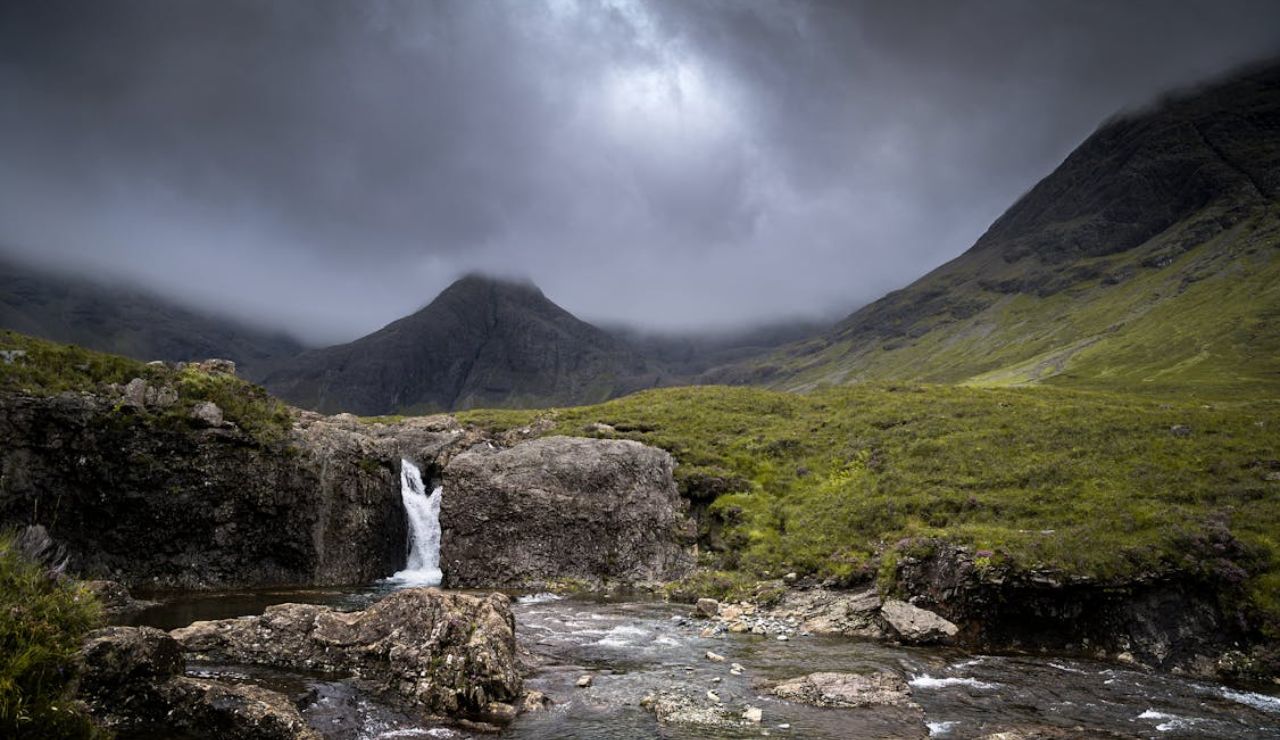 Dramatic cloudy sky over a rocky waterfall and lush green valley in the Fairy Pools of Isle of Skye, Scotland.