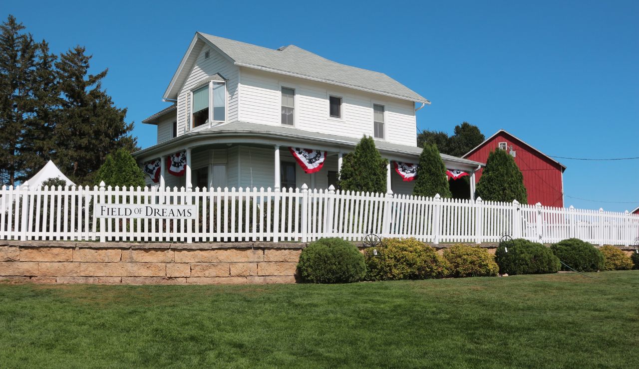 A white two-story farmhouse with a porch, adorned with red, white, and blue bunting, stands behind a "Field of Dreams" white picket fence.