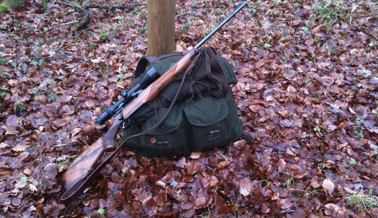 Hunting rifle with scope resting on a green backpack in a forest with fallen leaves.