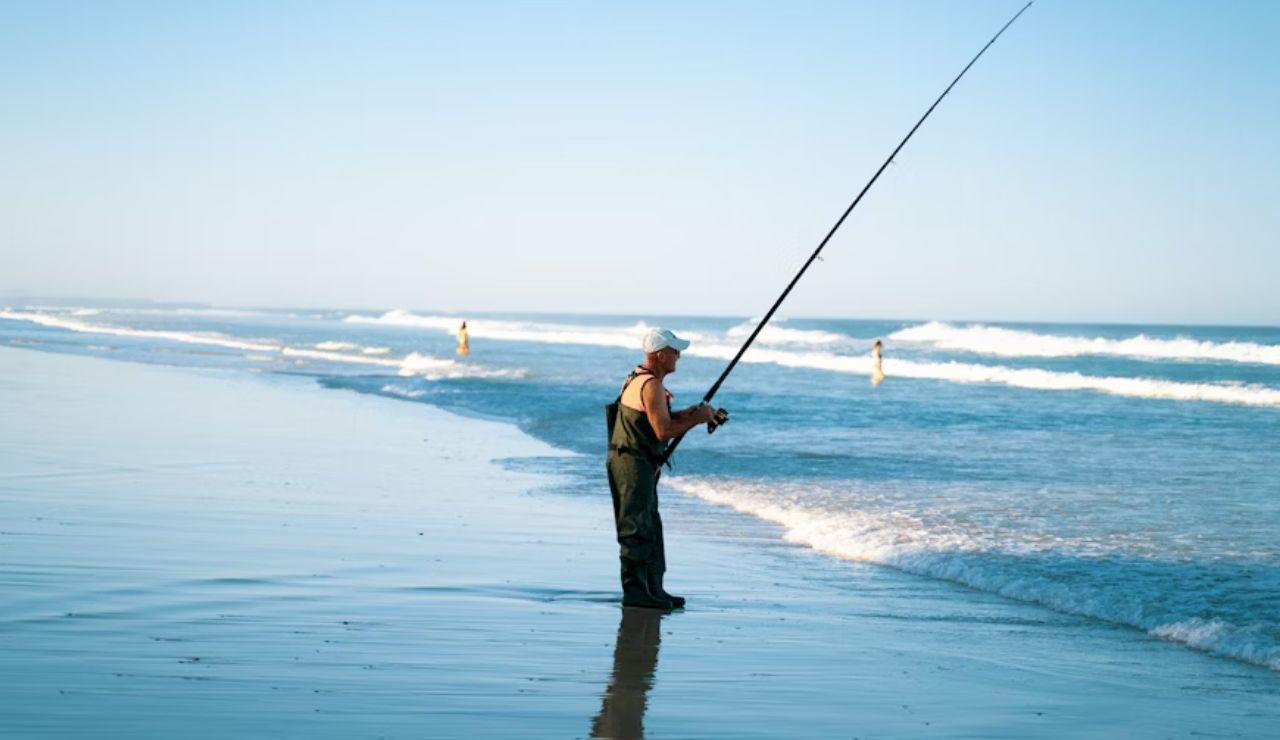Man wearing waders and a white cap fishing with a rod on a sandy beach by the ocean.