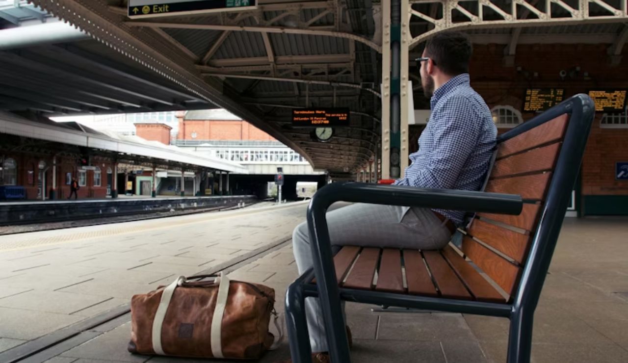 A man sitting on a bench at a train station platform with a brown leather duffel bag on the ground beside him, waiting for a train.