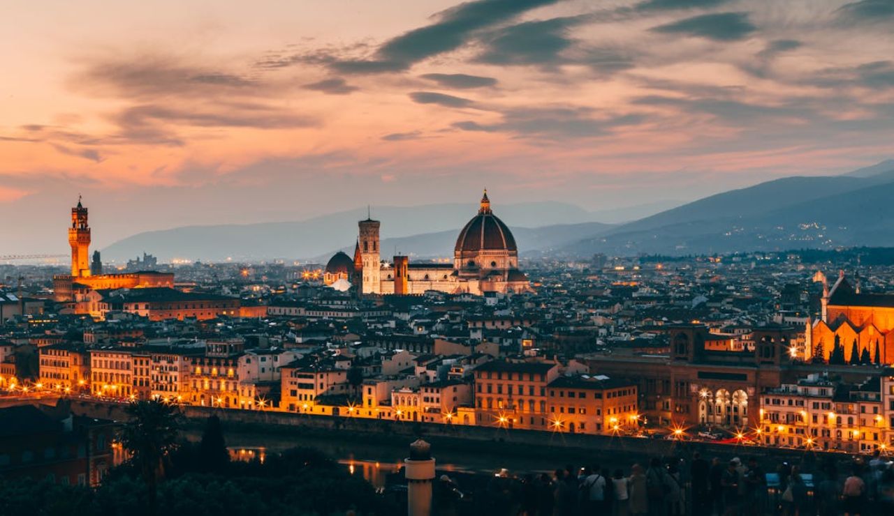 Panoramic view of Florence, Italy, at sunset with the illuminated Florence Cathedral (Duomo), Palazzo Vecchio, and city buildings under a colorful sky.