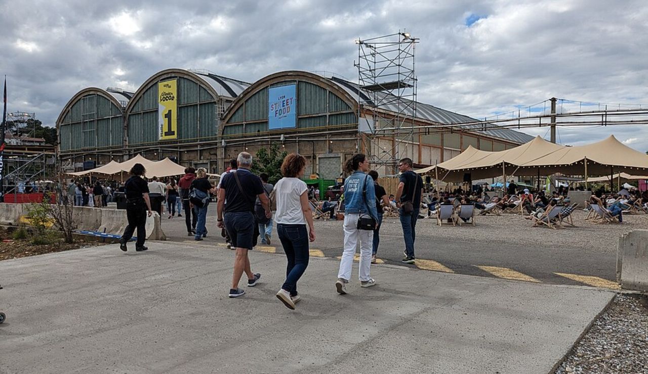 People walk toward a large industrial-style building hosting a street food festival, with outdoor tents, lounge chairs, and festive lighting.