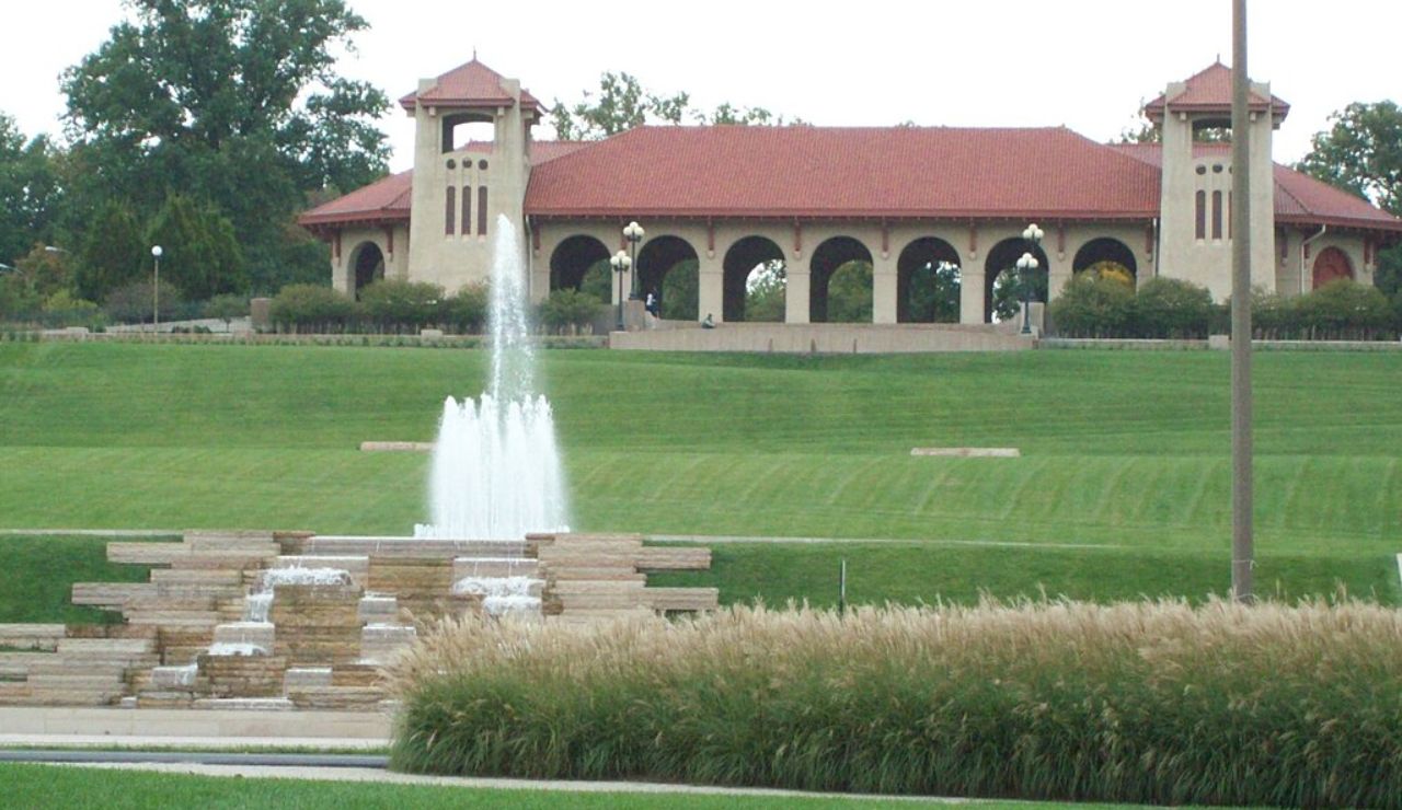 A pavilion with red-tiled roof and arched walkways sits atop a grassy hill, with a tiered water fountain and ornamental grass in the foreground.