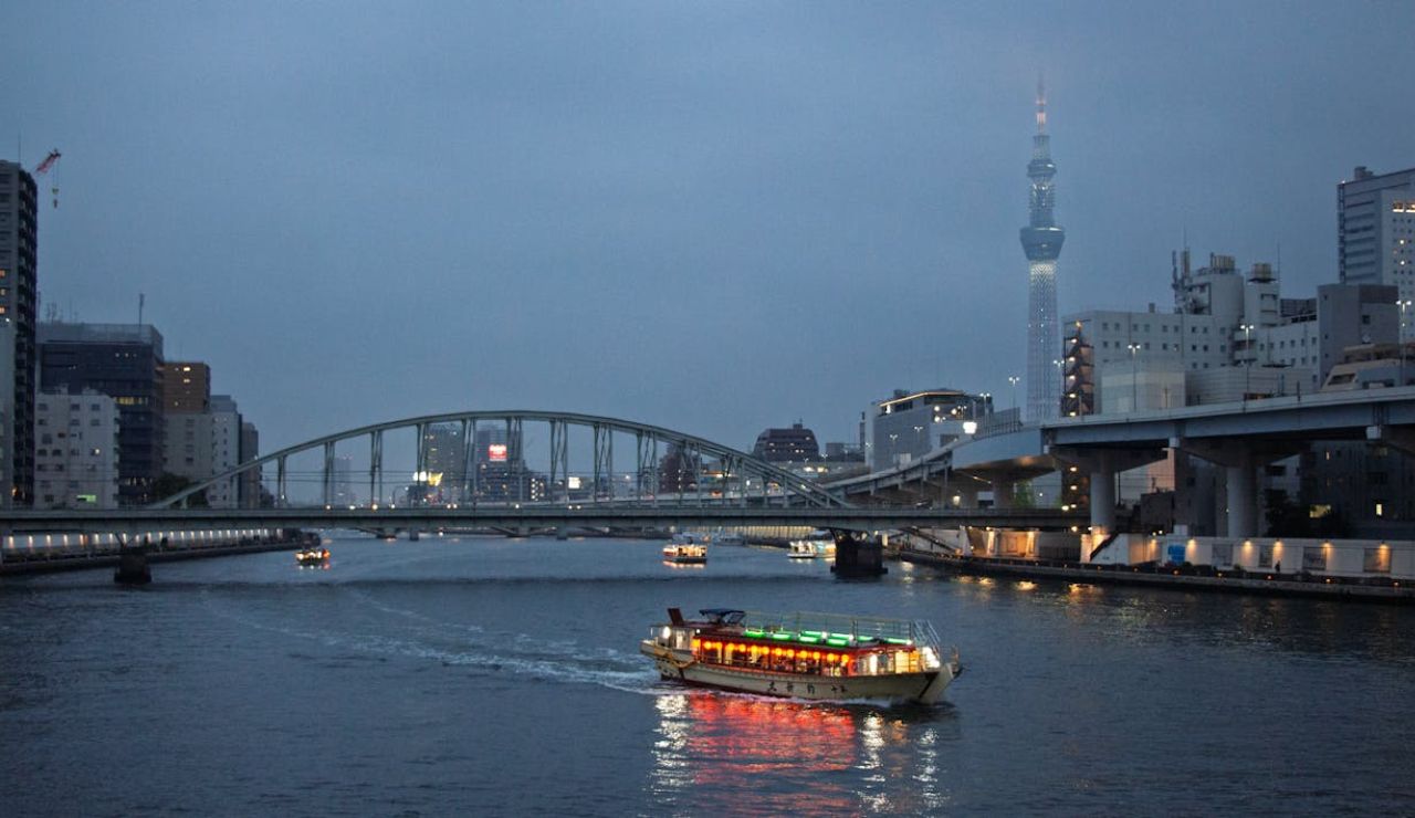 A brightly lit riverboat cruises along a city river at dusk in Tokyo, with the Tokyo Skytree and bridges visible in the background.