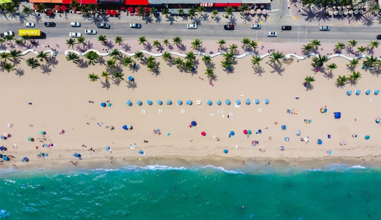 Aerial view of a busy beach with colorful umbrellas, palm trees, and a road lined with cars and buildings along the shoreline.