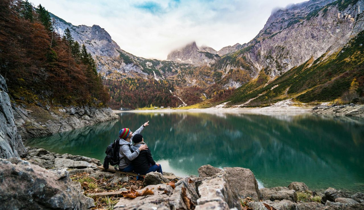 Two people sit on rocky ground by a tranquil mountain lake, with one pointing toward the misty peaks surrounded by autumn-colored forests.