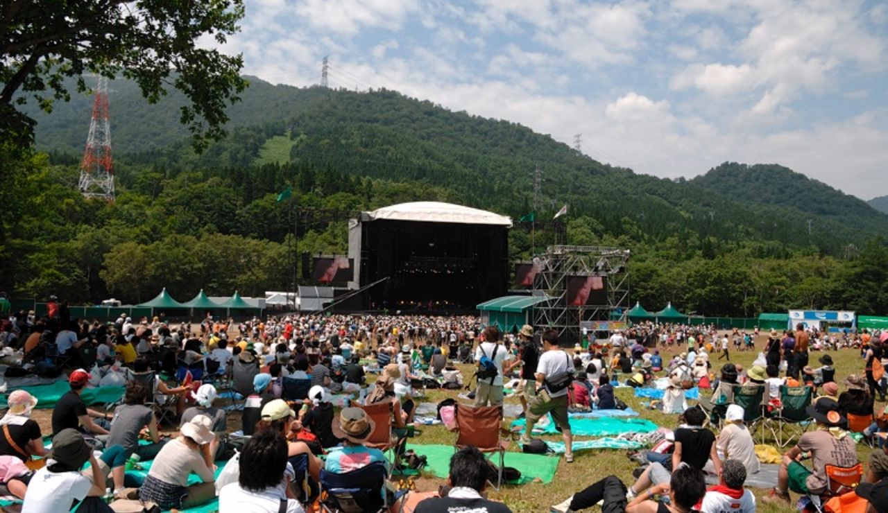 A large crowd gathered on a grassy field at an outdoor music festival, facing a stage set against a scenic backdrop of forested mountains.