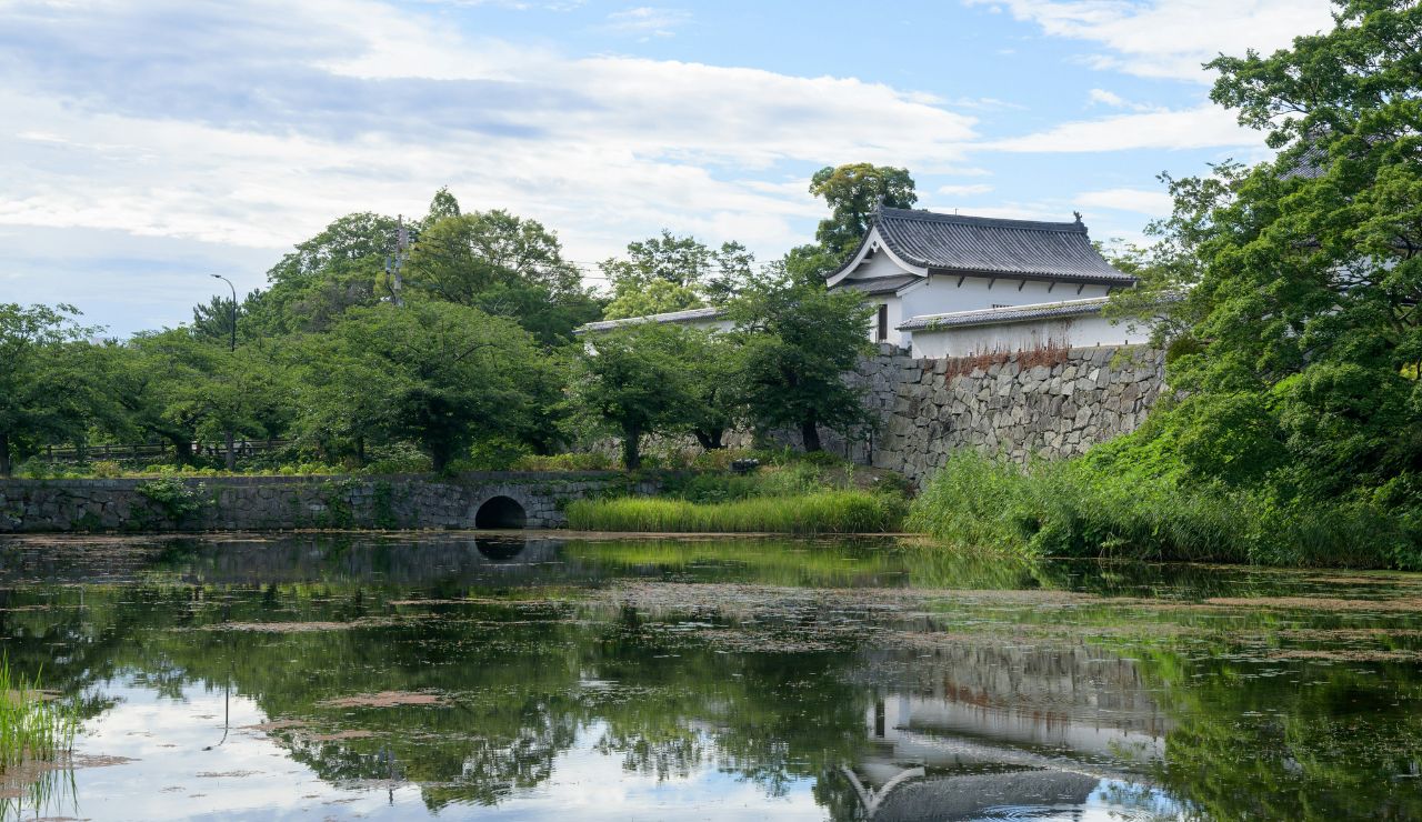 A traditional Japanese building with white walls and a black tiled roof stands beside a stone wall, surrounded by trees and a reflecting pond.