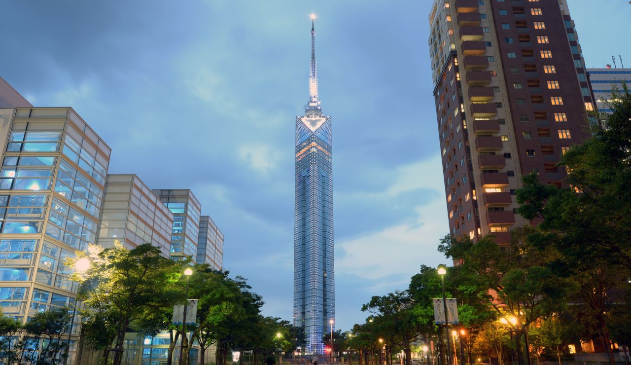 A tall, illuminated glass tower with a spire stands among modern buildings and trees under a cloudy dusk sky.