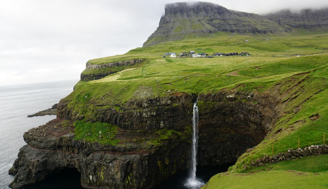 A waterfall cascades from a grassy cliff into the ocean, with a village and flat-topped mountain in the background beneath a cloudy sky.