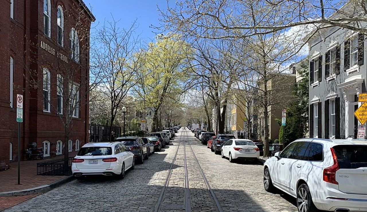 Cobbled city street lined with parked cars, early spring trees, and historic brick and townhouse buildings under a clear blue sky.