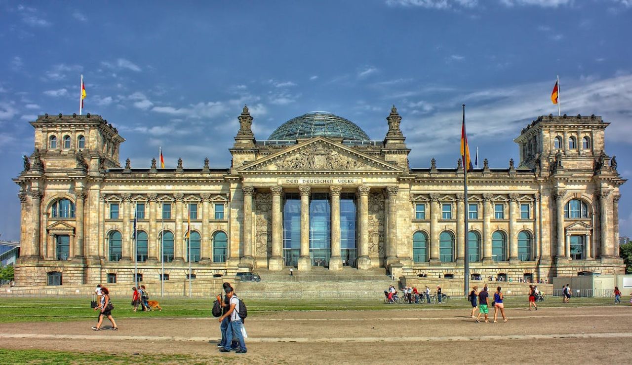 The Reichstag building in Berlin, Germany, featuring its grand classical architecture and iconic glass dome, with people walking in the foreground.