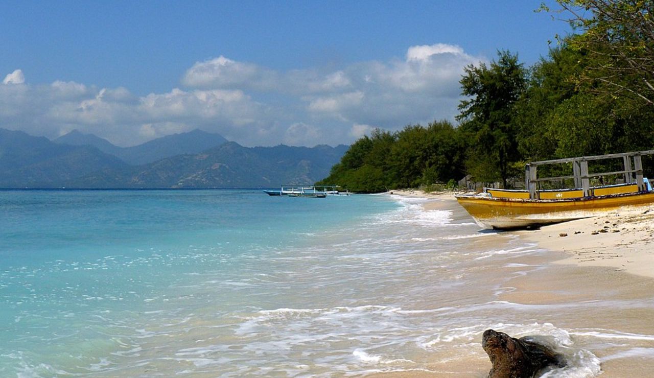 Tropical beach with calm waves, yellow boats on the shore, lush greenery, and distant mountains under a partly cloudy sky