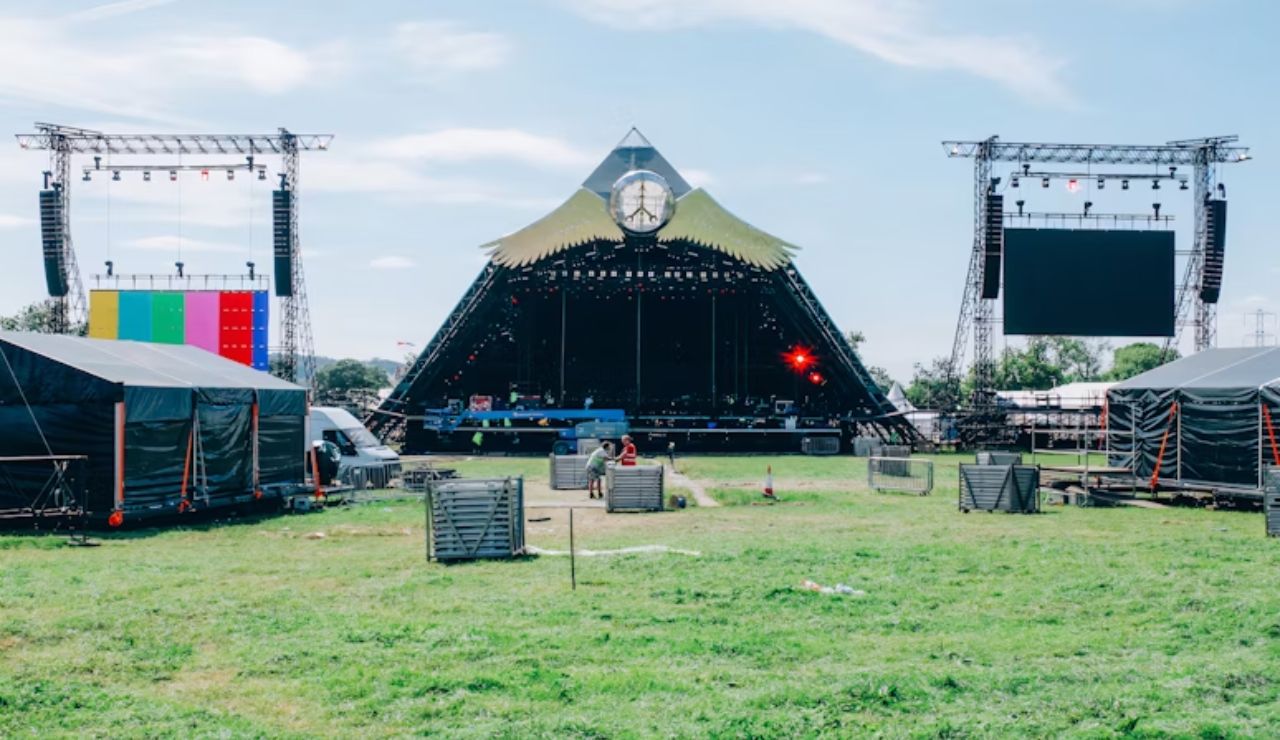 A large outdoor music stage under construction, with lighting rigs and speaker towers set up on a grassy field.