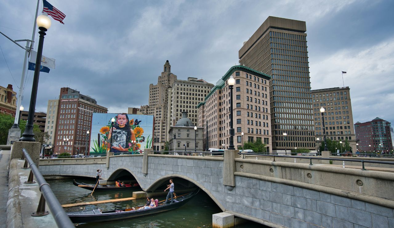 Urban canal scene in Providence, Rhode Island with gondolas under a bridge, a large mural, and tall buildings under a cloudy sky.