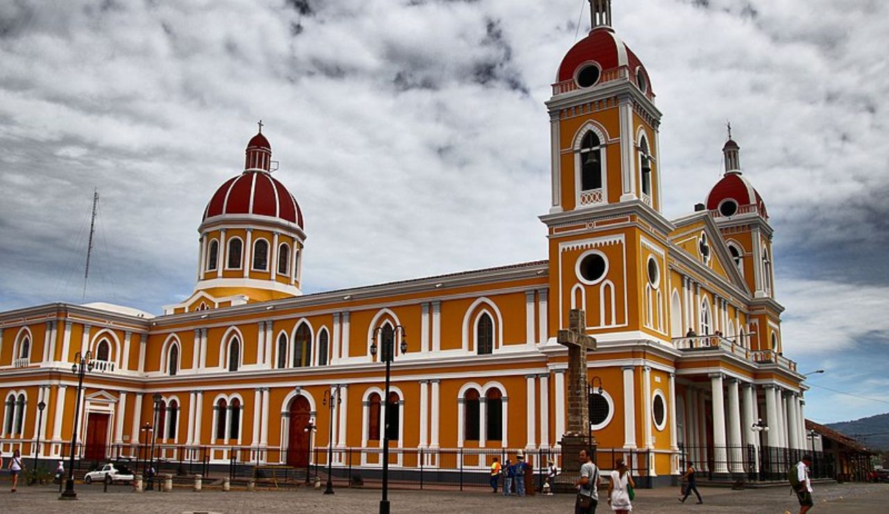 Vibrant yellow and white cathedral with red domes and arched windows in Granada, Nicaragua, set against a dramatic cloudy sky.