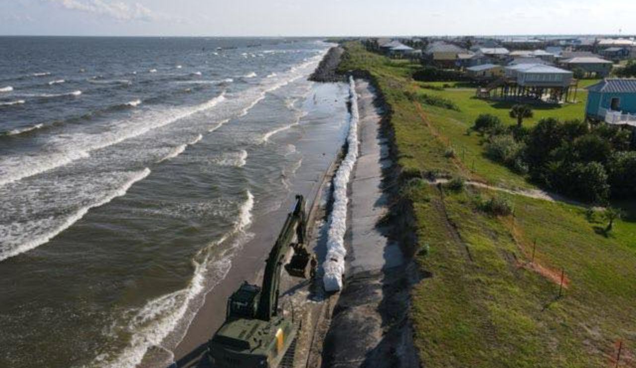 Excavator placing sandbags along a shoreline for coastal erosion control near beachfront homes.