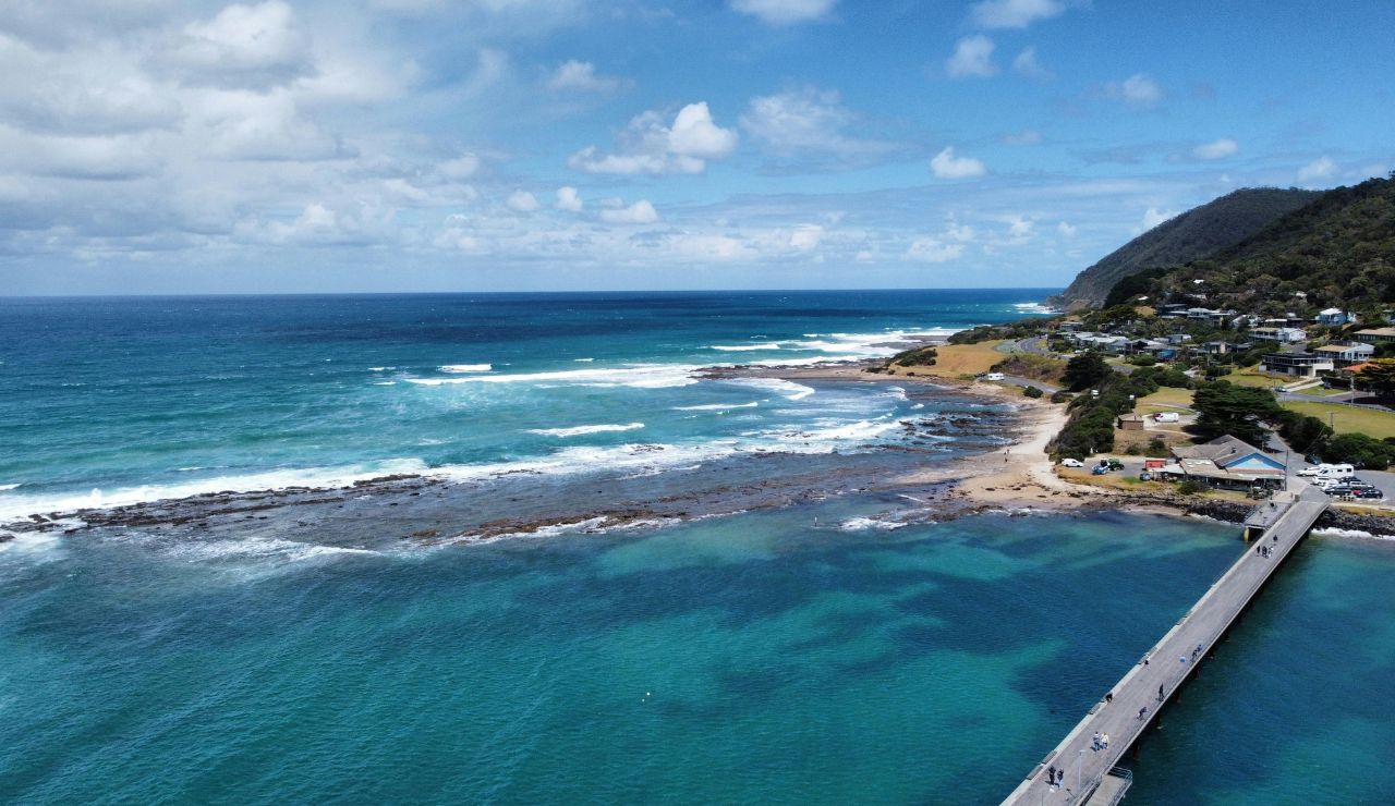 Aerial view of a coastal pier stretching into vibrant turquoise waters, with waves crashing on rocky shores and a lush green hillside town in the background.