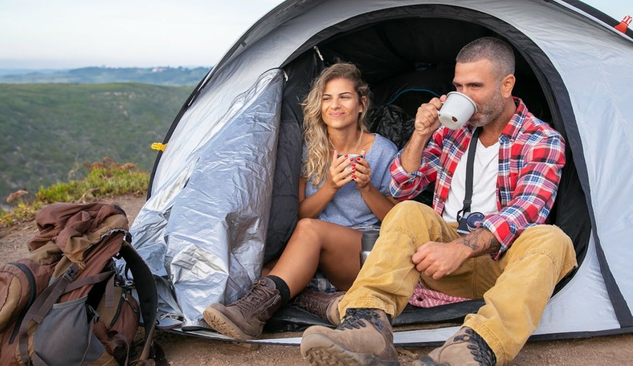 A smiling couple sits at the entrance of a tent on a mountaintop, enjoying hot drinks with backpacks beside them and scenic hills in the background.