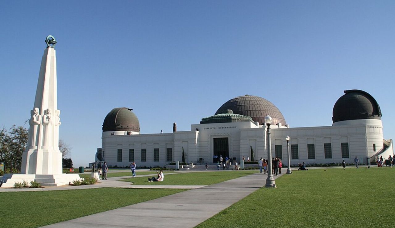 Griffith Observatory with its domed structure and white facade, set on a grassy lawn under a clear blue sky, with a monument in the foreground.