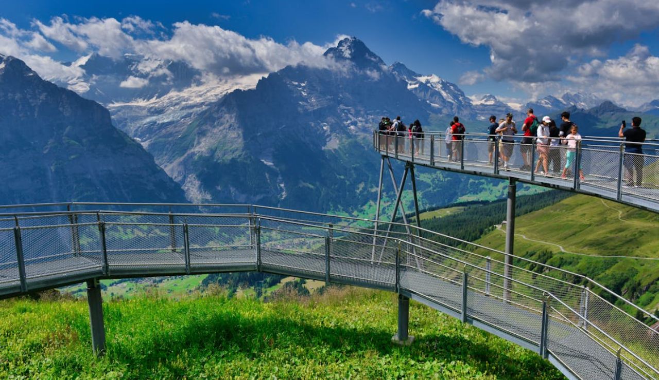 Tourists stand on a scenic metal walkway overlooking a breathtaking panorama of green valleys and snow-capped mountains under a partly cloudy sky.