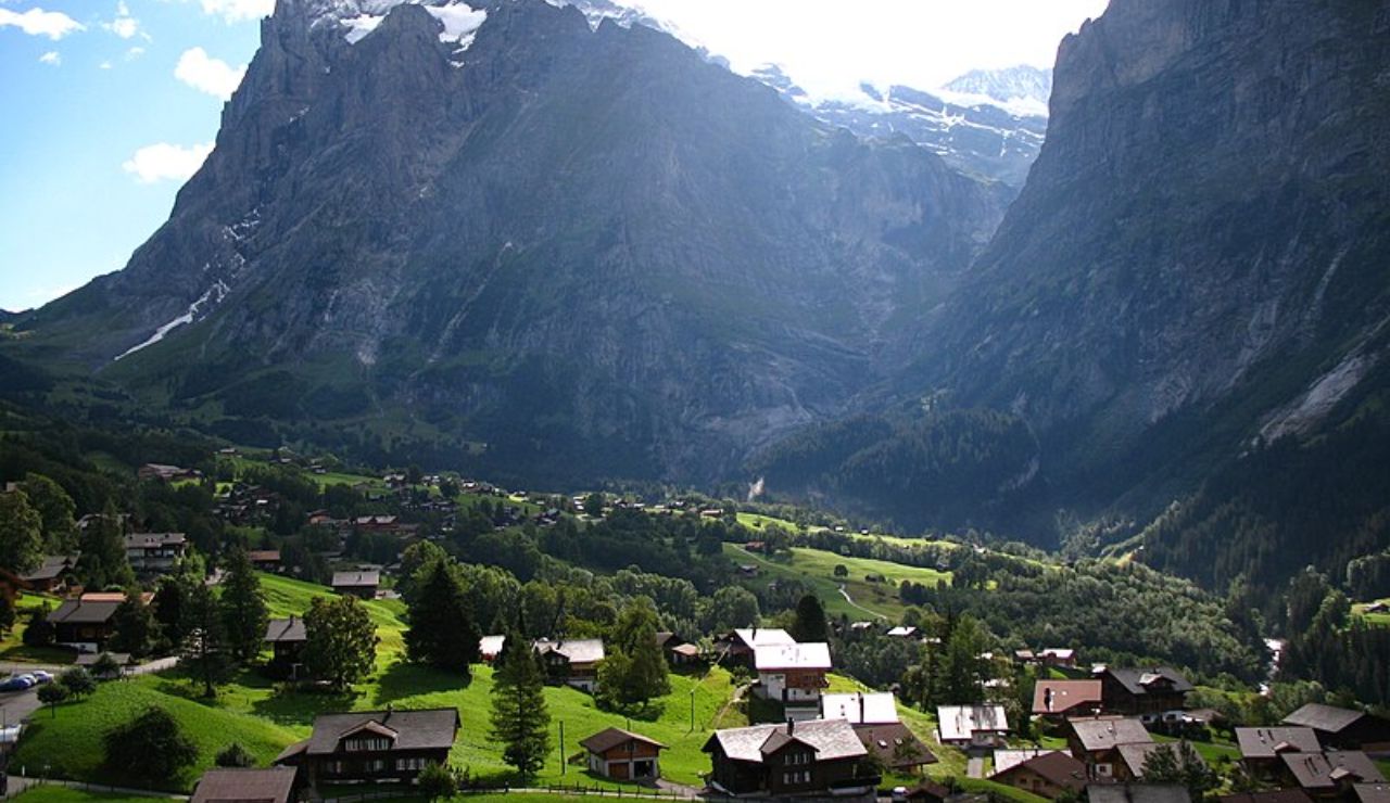 Charming alpine village with wooden houses nestled in a lush green valley, surrounded by towering rocky mountains and dappled sunlight.