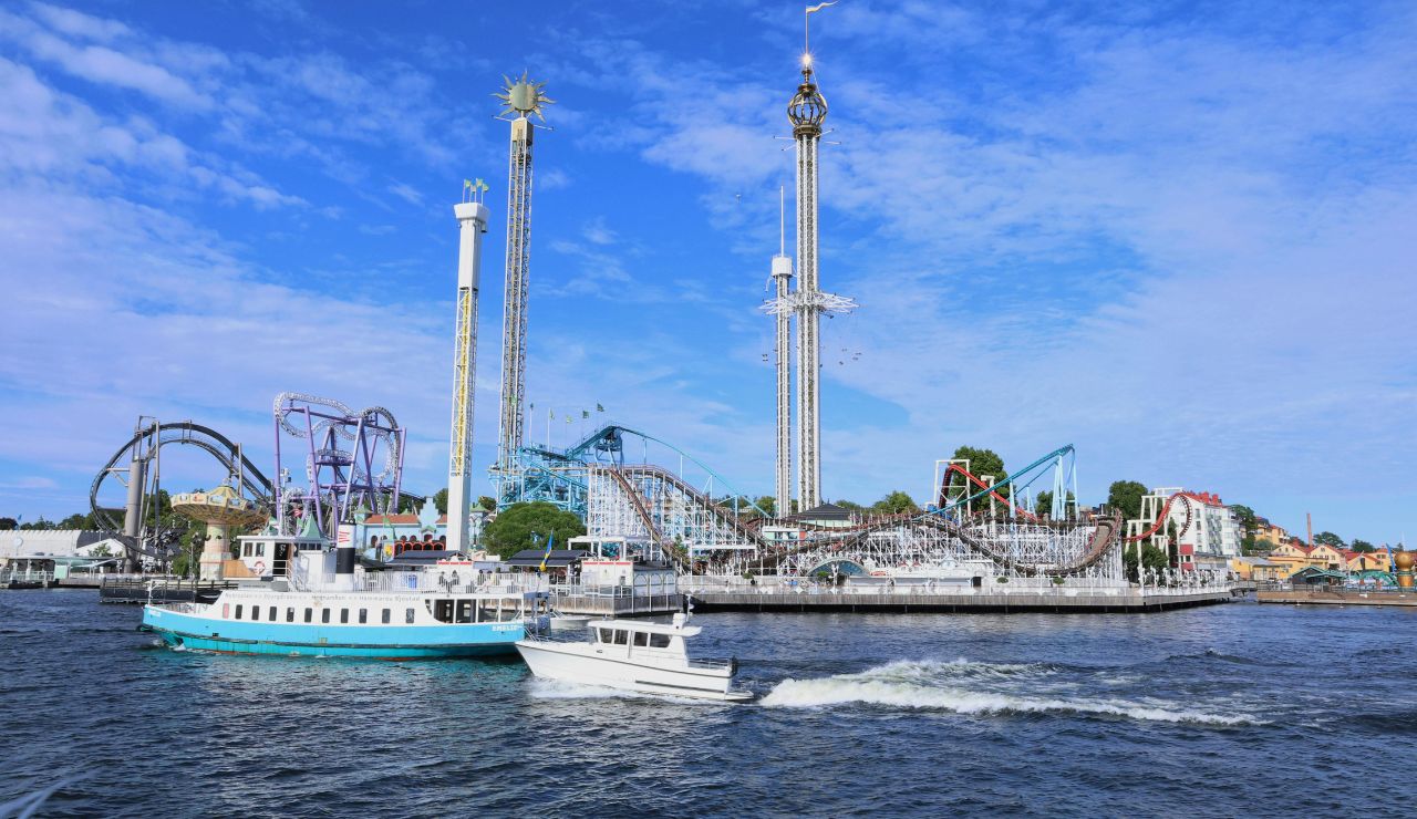 Boats passing by Gröna Lund amusement park in Stockholm, Sweden, with roller coasters and tall thrill rides visible under a bright blue sky.