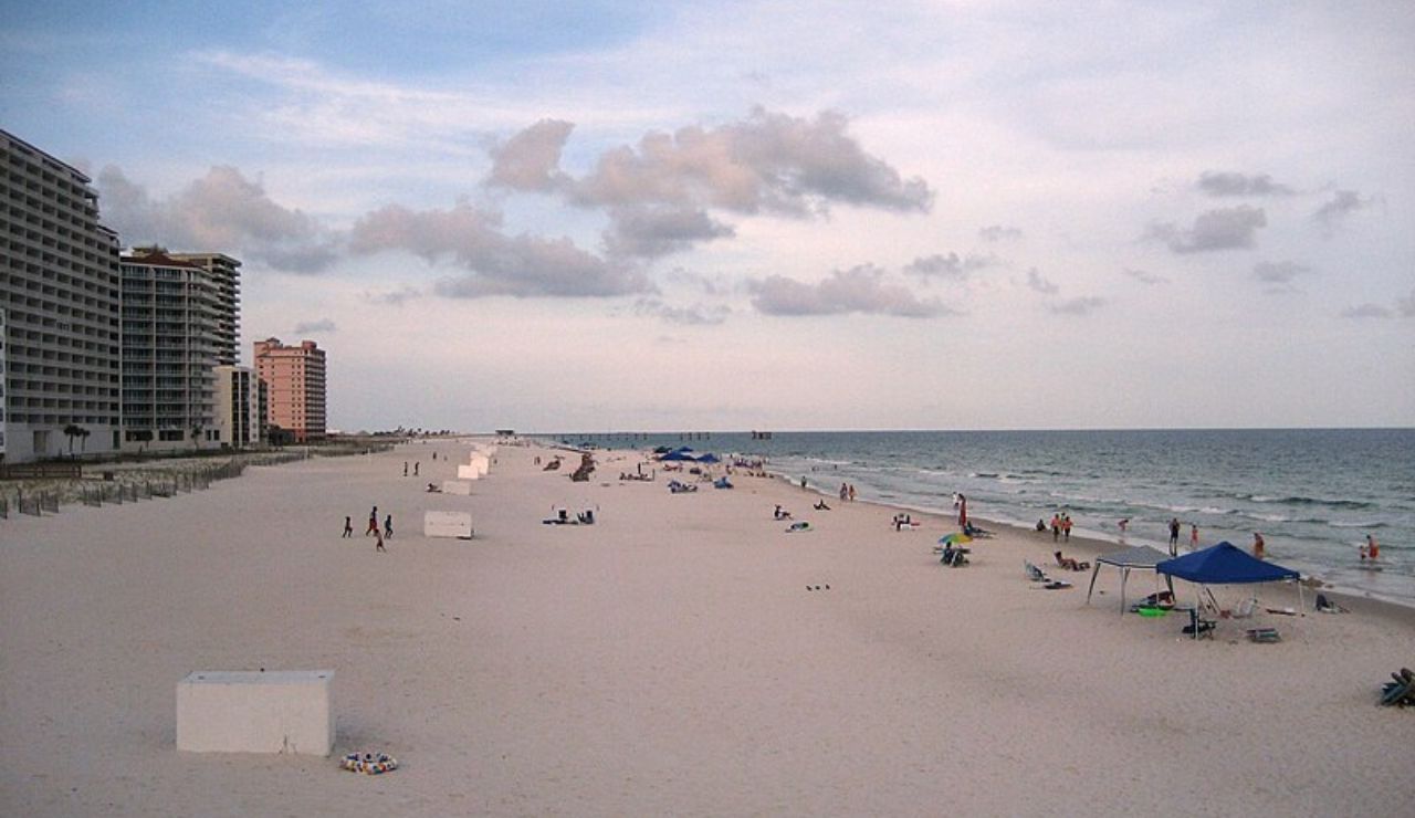 Wide sandy beach with scattered people, beach umbrellas, and high-rise buildings along the shoreline under a cloudy sky.